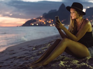 Young woman sitting at beach at night, looking at her mobile phone